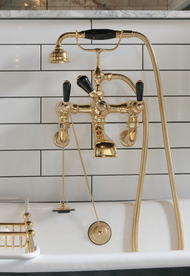 Glistening gold bath taps with black ceramic handles are wall mounted on white tiles with black grout above roll top bath.